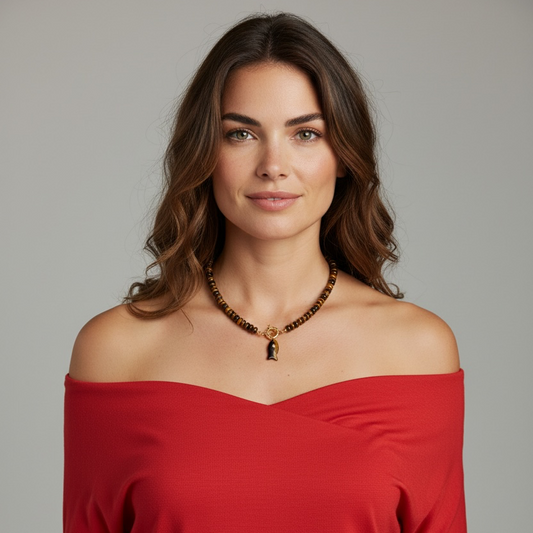 A woman with wavy brown hair wearing a red off-the-shoulder top and a beaded necklace with a small pendant, photographed against a plain light gray studio background.