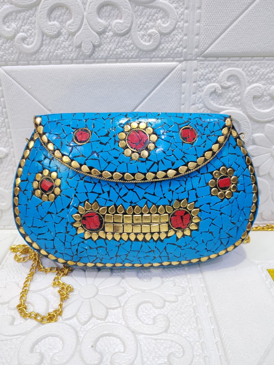 A small turquoise blue mosaic handbag with gold-tone metal embellishments, red gemstone accents, and a gold chain strap, displayed against a white textured background.