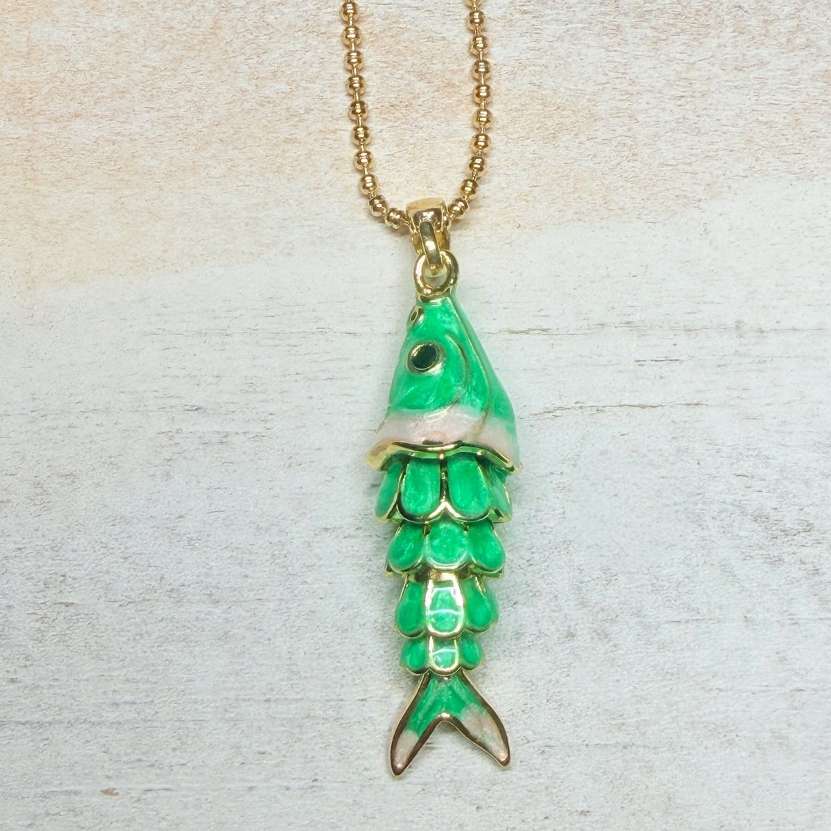 A gold chain necklace with a fish-shaped pendant featuring green and white enamel detailing, displayed on a light textured background.