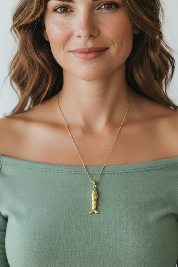 A woman wearing a delicate gold chain necklace with a small fish-shaped pendant, styled with a sage green off-the-shoulder top against a light gray background.
