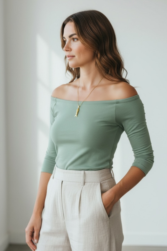 A woman wearing a sage green off-the-shoulder long-sleeve top and light beige high-waisted trousers, standing in a bright, minimalist room.