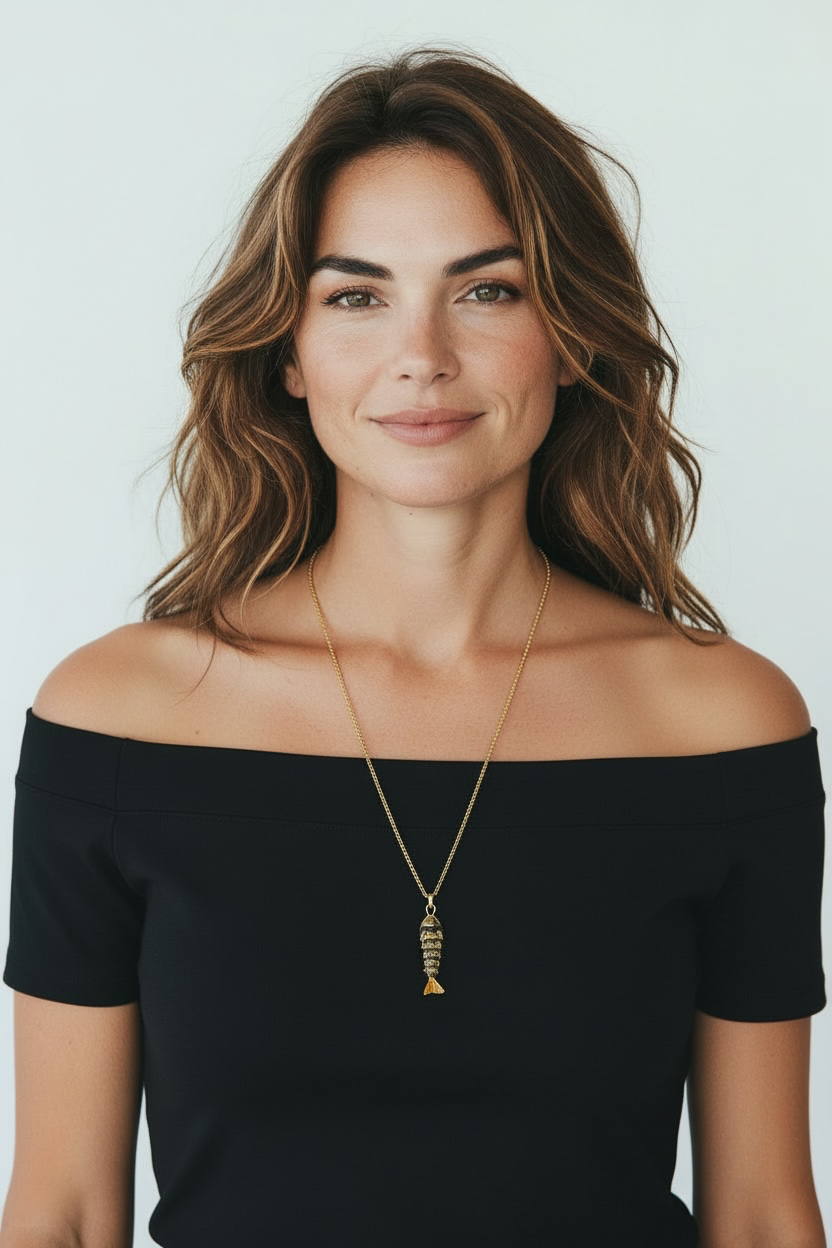 A woman with wavy brown hair wearing a black off-the-shoulder top and a gold pendant necklace with a fish-shaped charm, photographed against a light gray background.