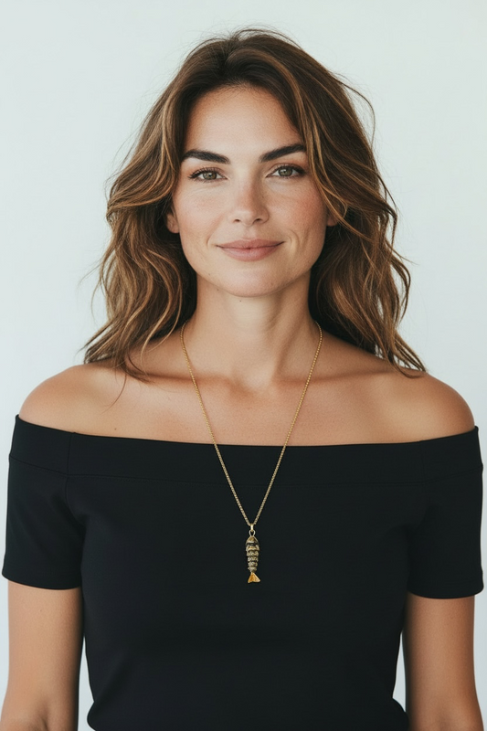 A woman with wavy brown hair wearing a black off-the-shoulder top and a gold pendant necklace with a fish-shaped charm, photographed against a light gray background.