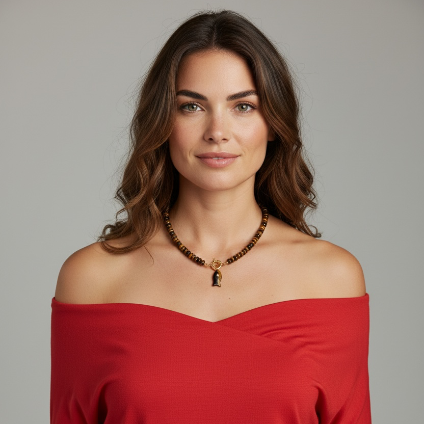 A woman with wavy brown hair wearing a red off-the-shoulder top and a beaded necklace with a small pendant, photographed against a plain light gray studio background.