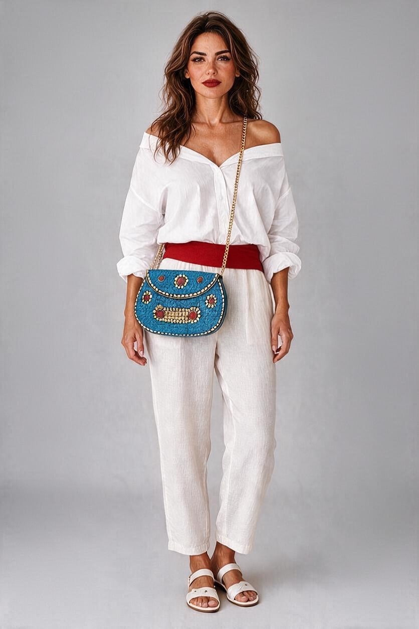 A woman modeling a white off-the-shoulder blouse and white wide-leg trousers, styled with a red belt and a blue beaded crossbody bag with gold chain straps, against a solid gray studio background.